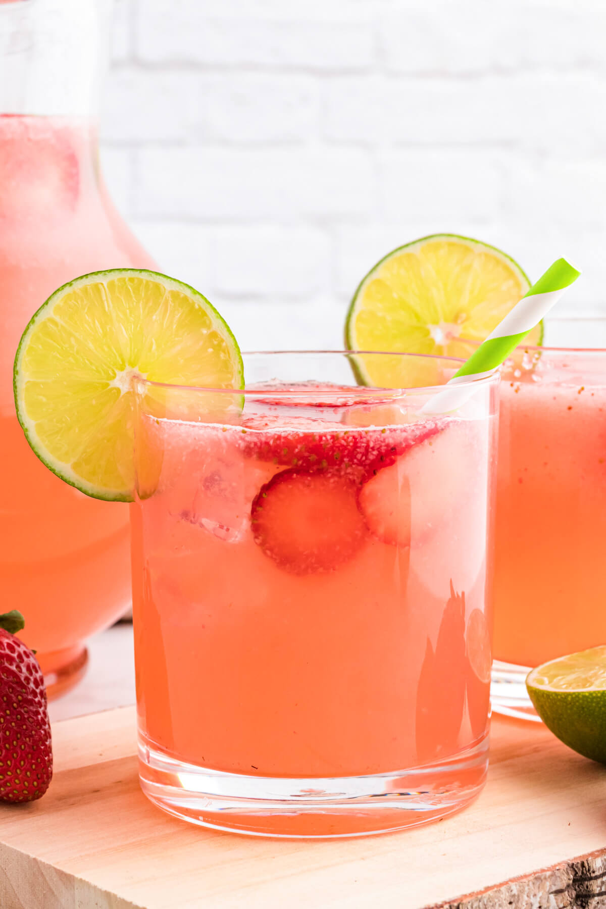A glass pitcher and glass full of pink Strawberry Limeade garnished with sliced strawberries and lime wheels.