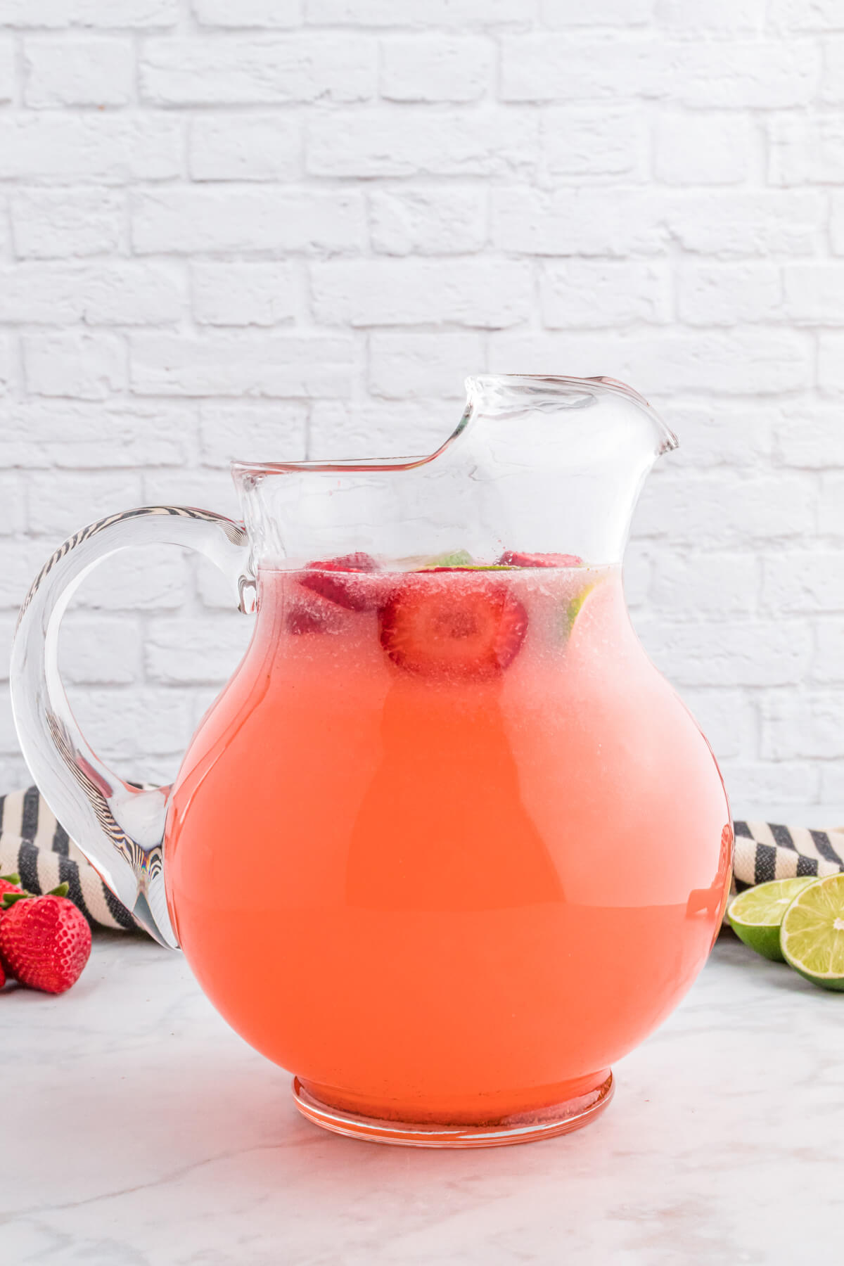 A glass pitcher filled with pink Strawberry Limeade garnished with sliced strawberries and lime wheels.