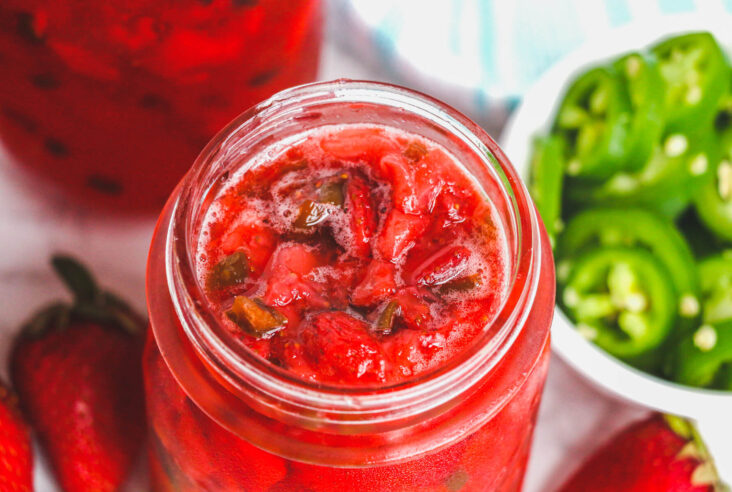 An open jar of ruby red strawberry jalapeño jam surrounded by raw ingredients.