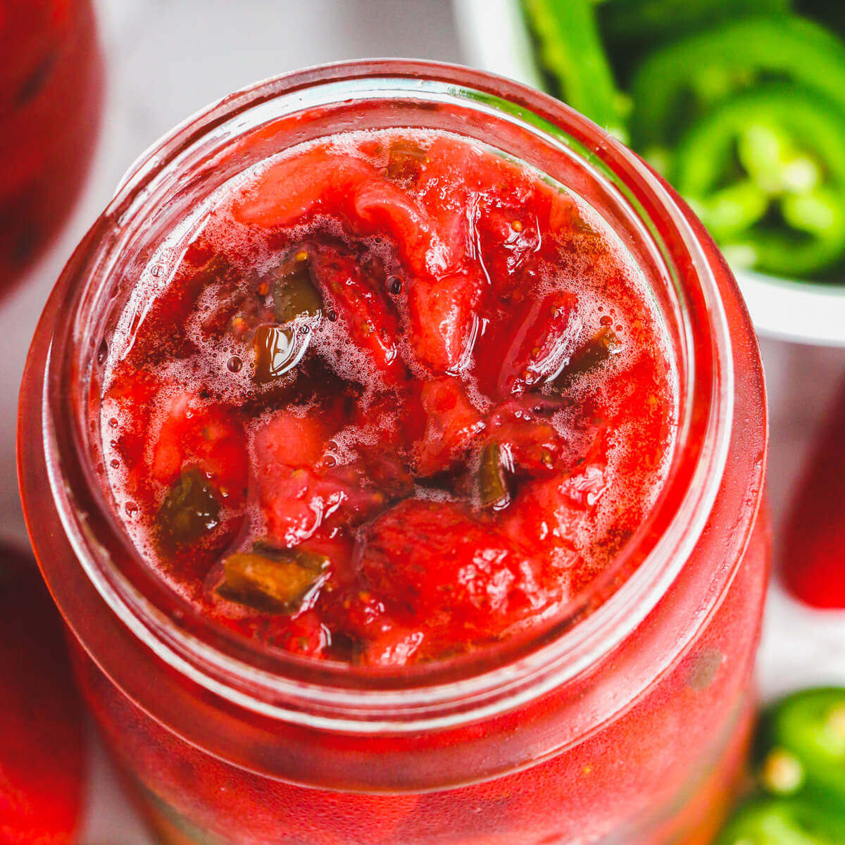 An open jar of ruby red strawberry jalapeño jam surrounded by raw ingredients.