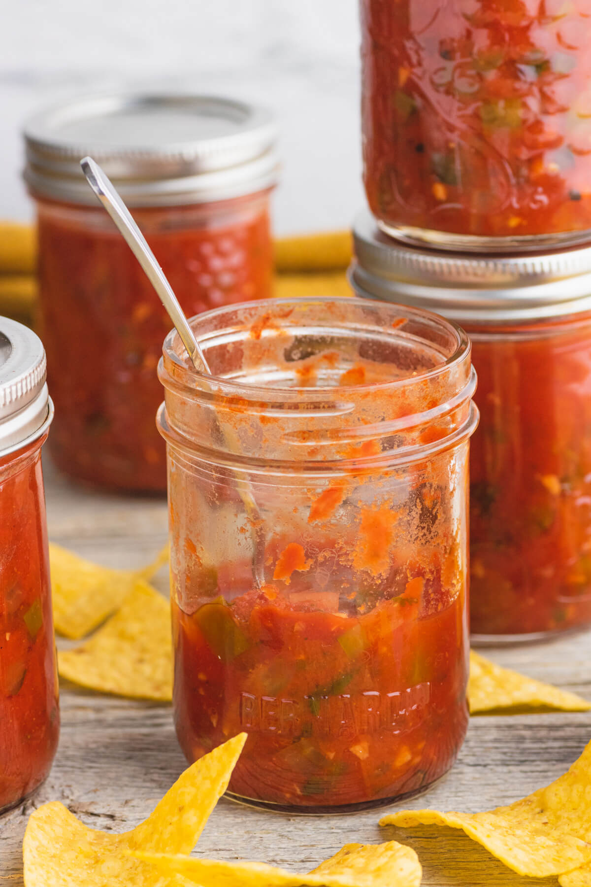 A half empty jar of salsa with a spoon in it surrounded by jars of canned salsa and tortilla chips.
