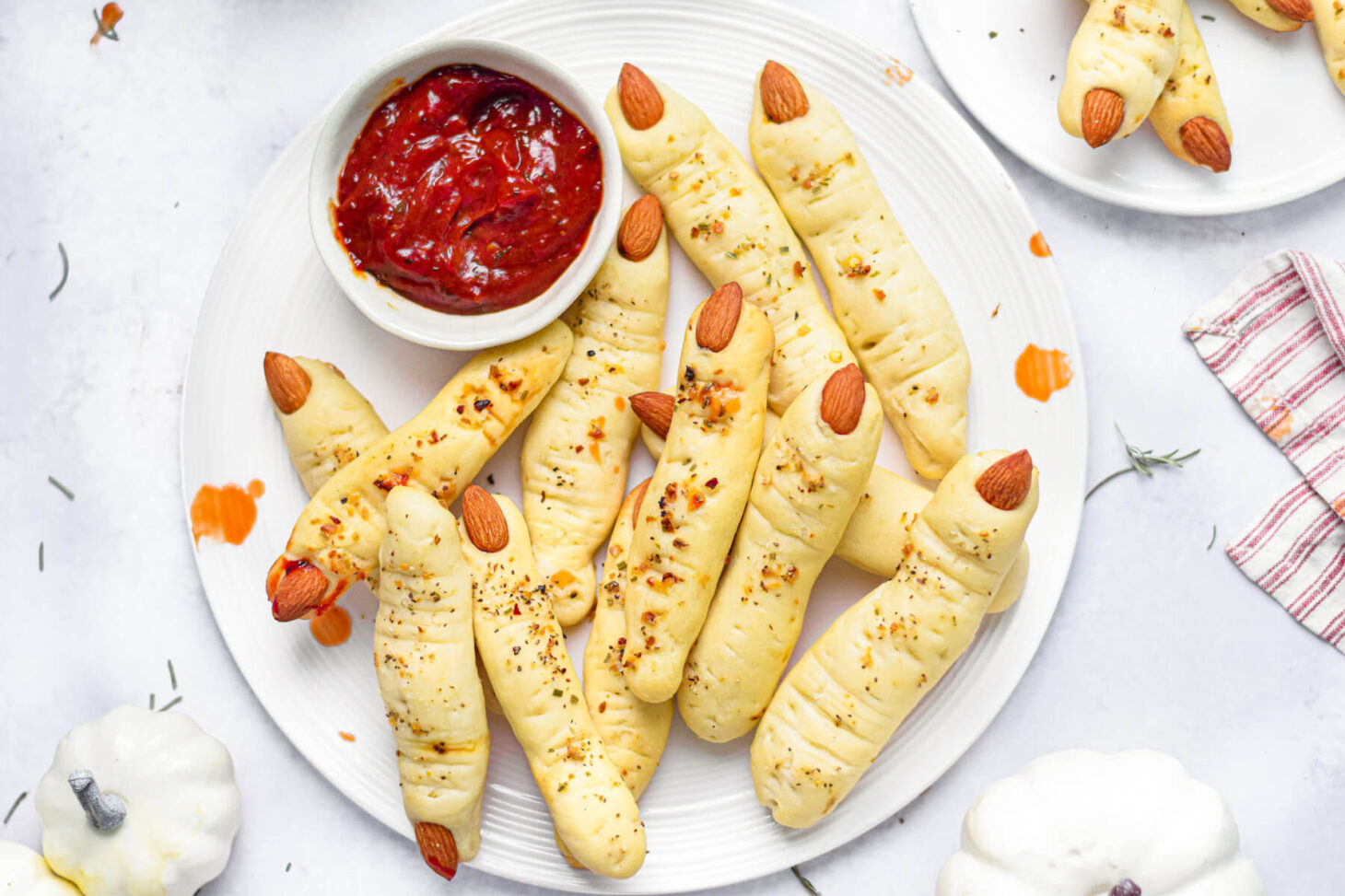 A white plate containing many creepy witch finger Italian breadsticks with a small bowl of marinara sauce for dipping nearby.