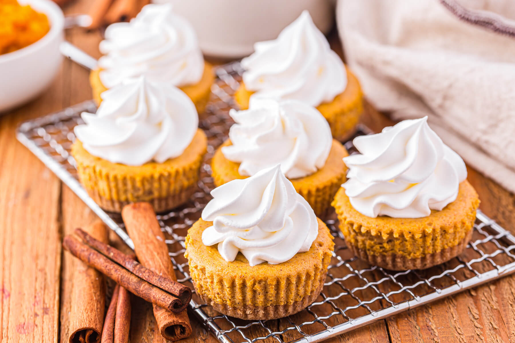 A wire baking rack containing six mini pumpkin cheesecakes topped with whipped cream.