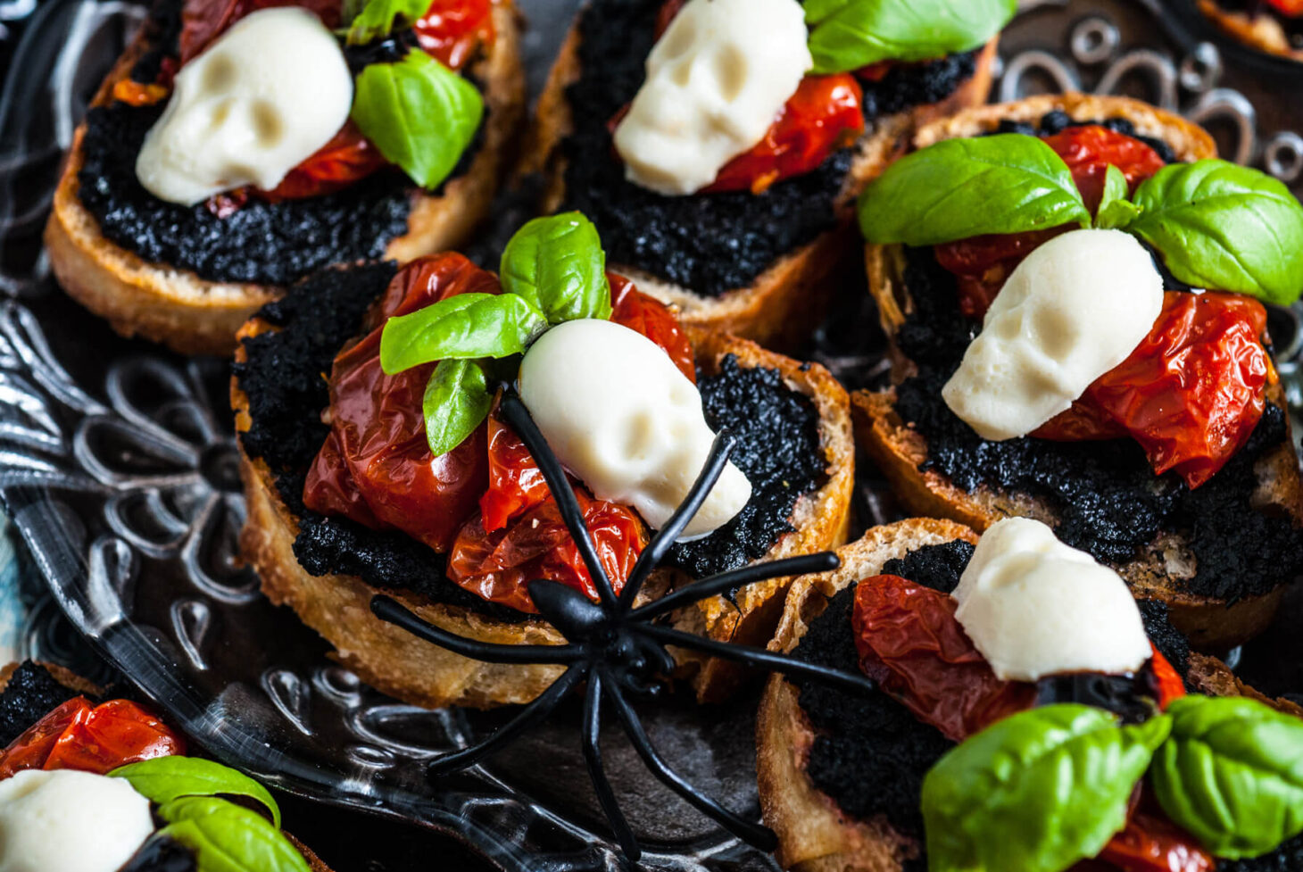 A spooky scene featuring a silver tray of caprese crostini decorated with mozzarella skulls and spiders.
