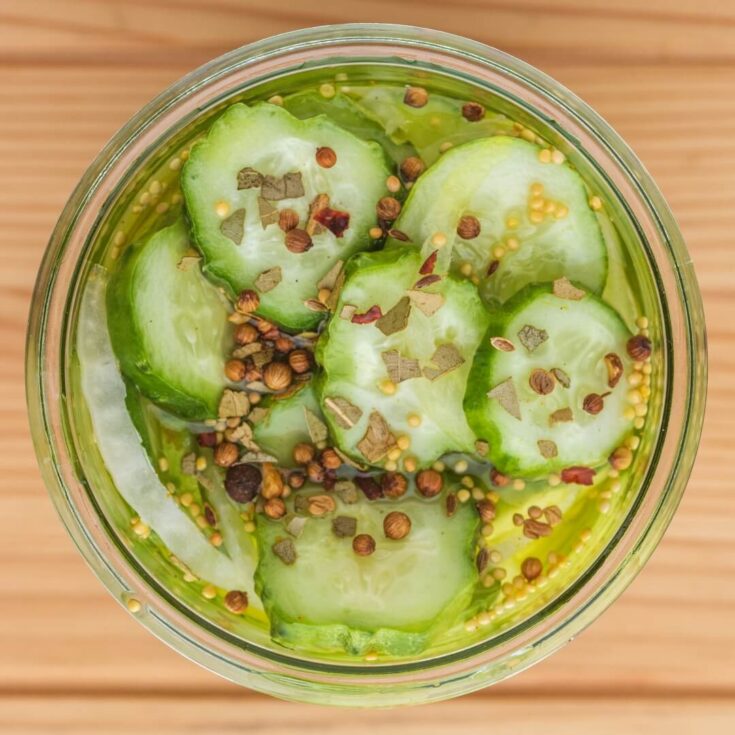A glass a jar filled with sliced cucumbers and onions topped with pickling spice and brine.
