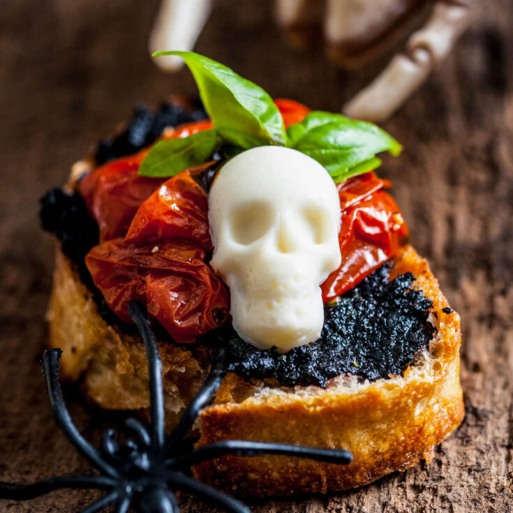A single spooky skull caprese crostini topped with black pesto, roasted tomatoes, a mozzarella skull, and fresh basil leaves.