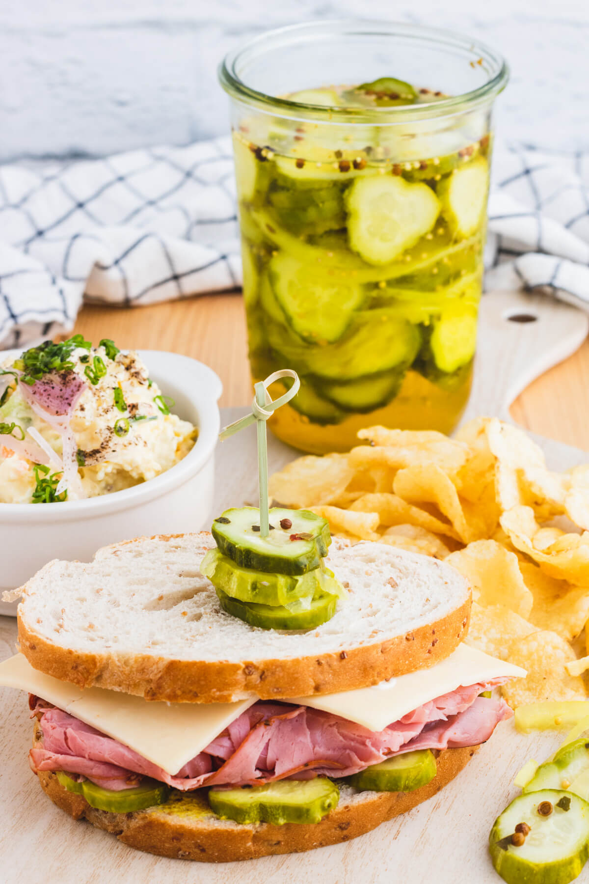 A table set with a jar of Refrigerator Bread and Butter pickles, potato salad, potato chips, and a sandwich garnished with sliced pickles.