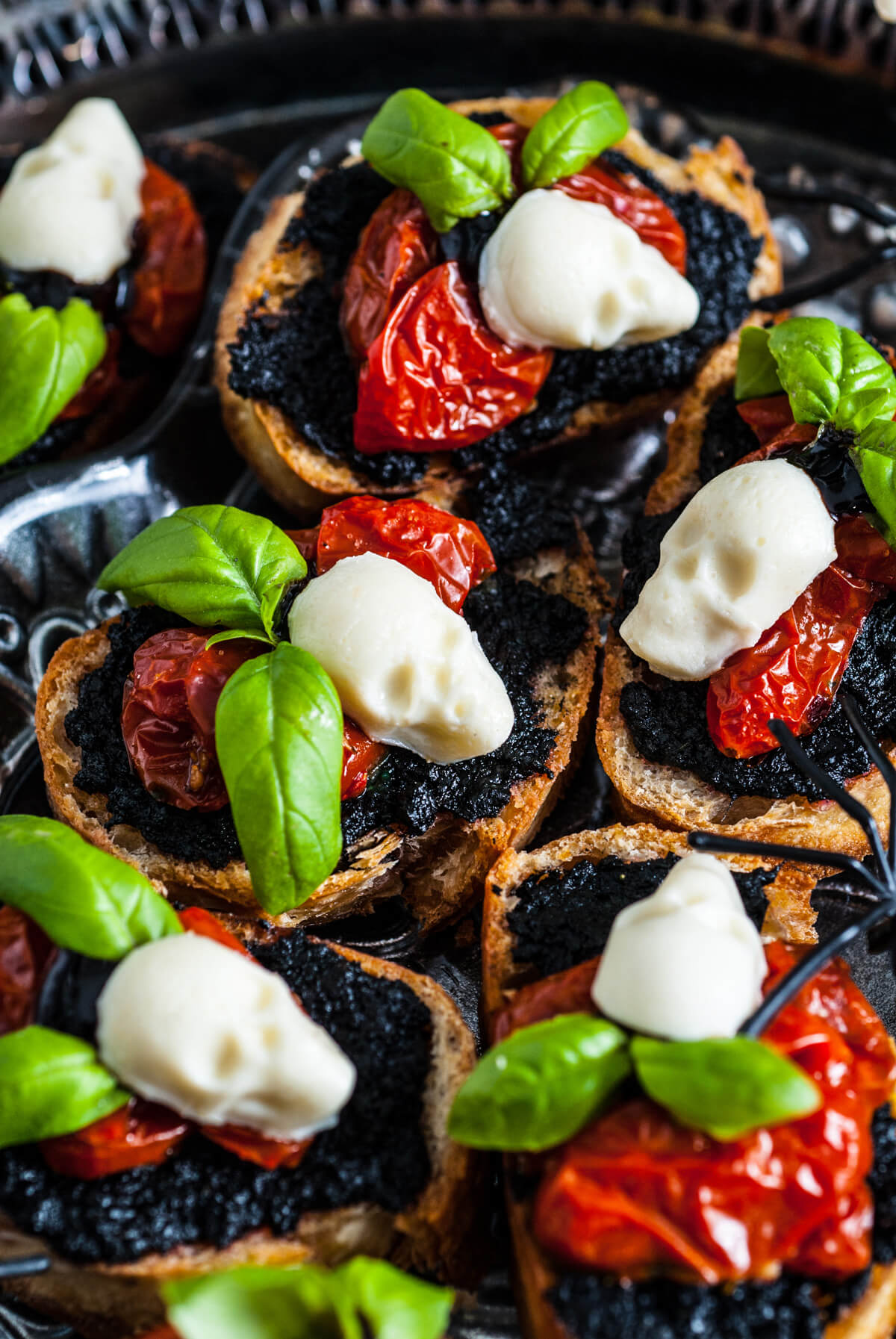 A platter of toasted crostini topped with black pesto, roasted cherry tomatoes, fresh basil leaves, and mozzarella ball skulls 