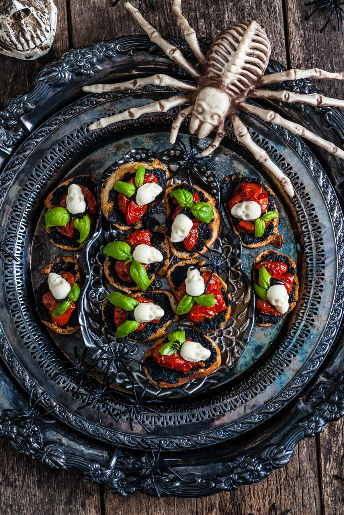 A spooky scene featuring a silver tray of caprese crostini decorated with mozzarella skulls and spiders. 

