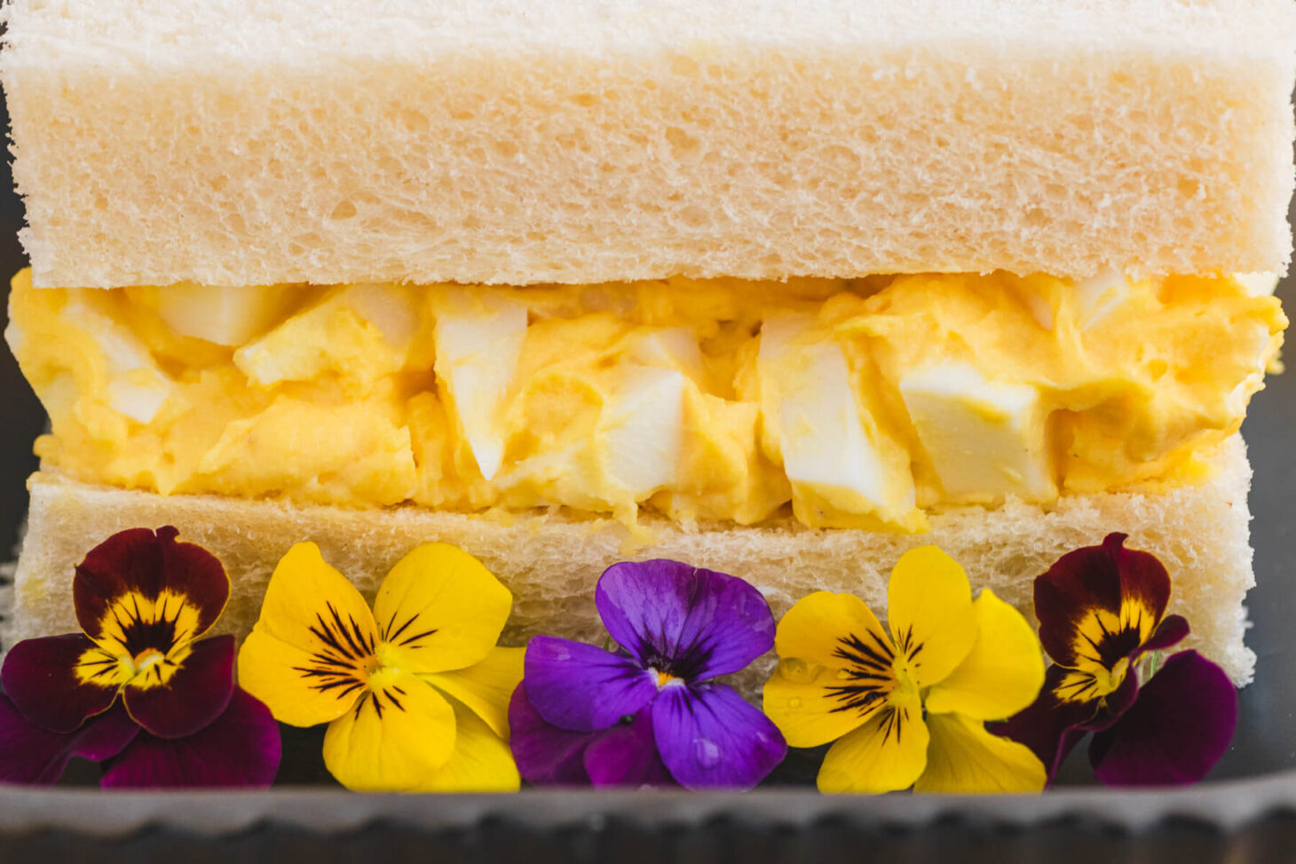 A rich yellow egg salad sandwich on fluffy white crust less bread on a plate garnished with edible flowers.