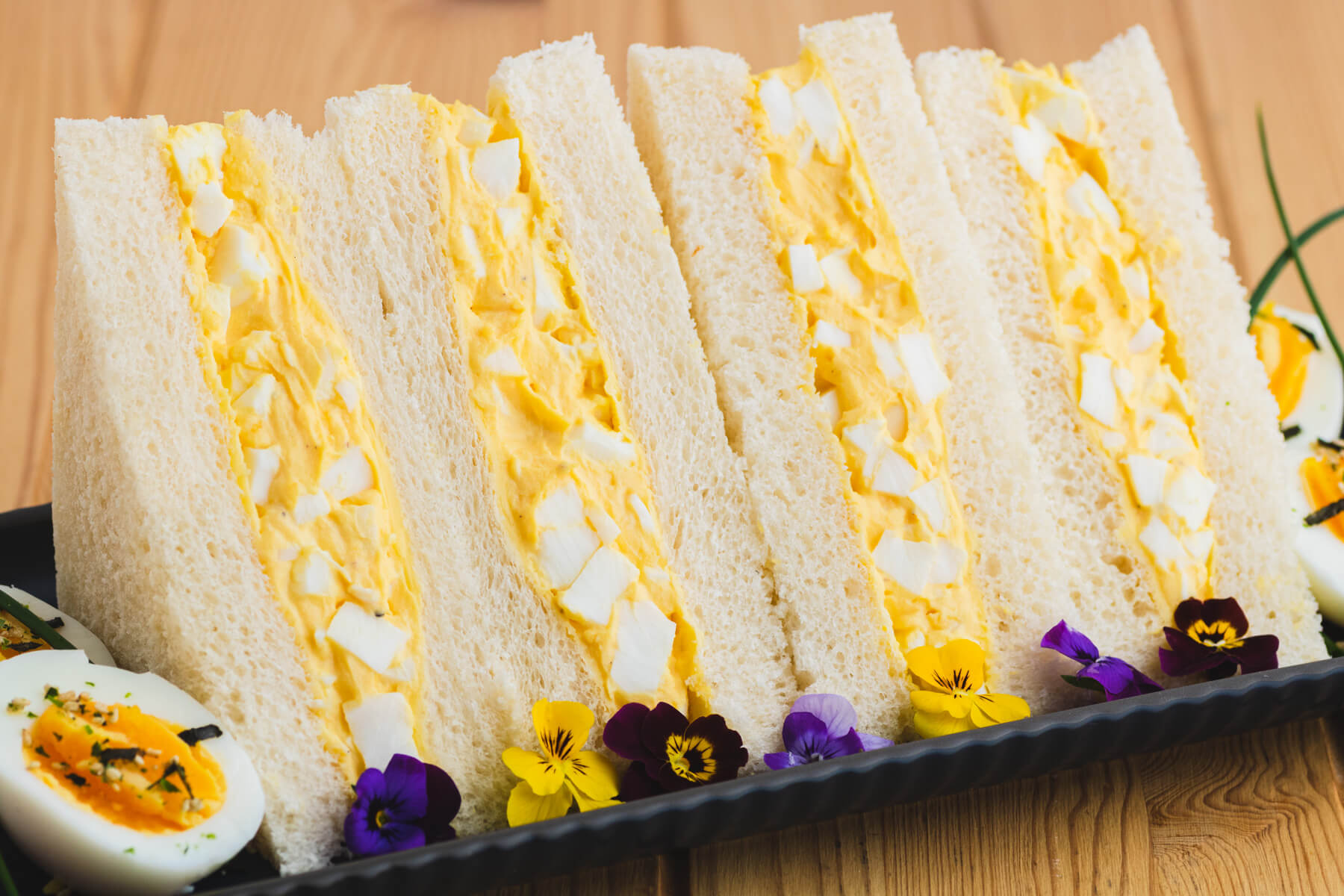 Four rich yellow egg salad sandwiches on fluffy white crust less bread on a plate garnished with a hard boiled egg and edible flowers.
