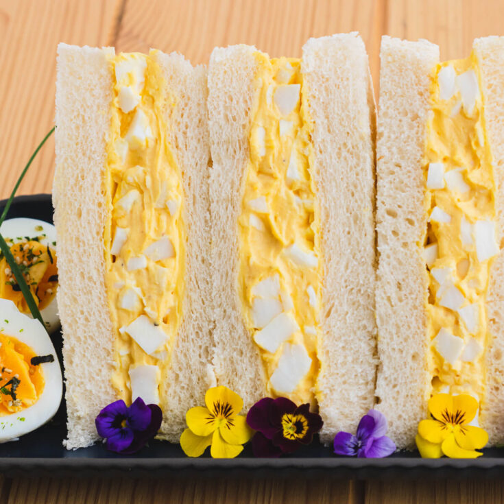 Three rich yellow egg salad sandwiches on fluffy white crust less bread on a plate garnished with a hard boiled egg and edible flowers.