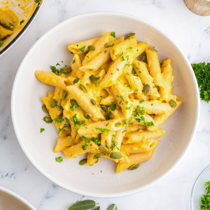 A white bowl containing creamy penne in a pumpkin pasta sauce garnished with pepitas and parsley.