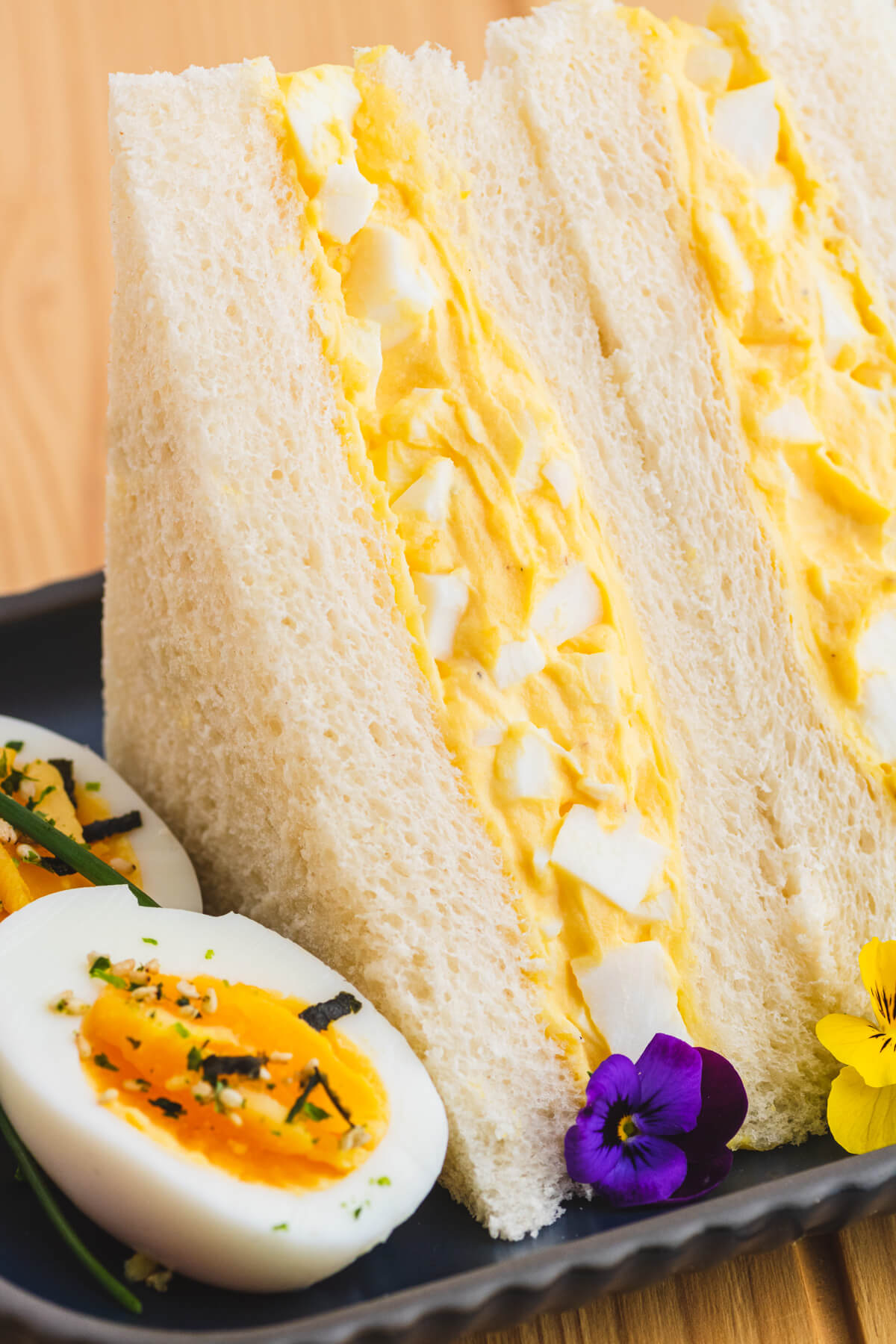 Two rich yellow egg salad sandwiches on fluffy white crust less bread on a plate garnished with a hard boiled egg and edible flowers.
