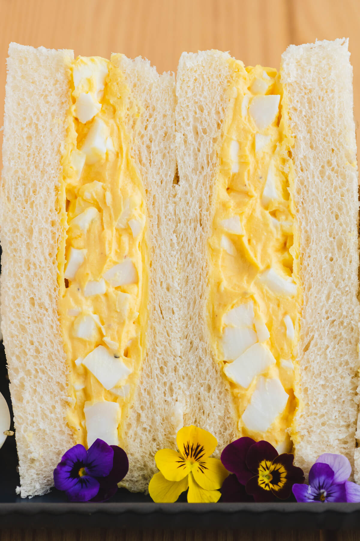 Two rich yellow egg salad sandwiches on fluffy white crust less bread on a plate garnished with edible flowers.