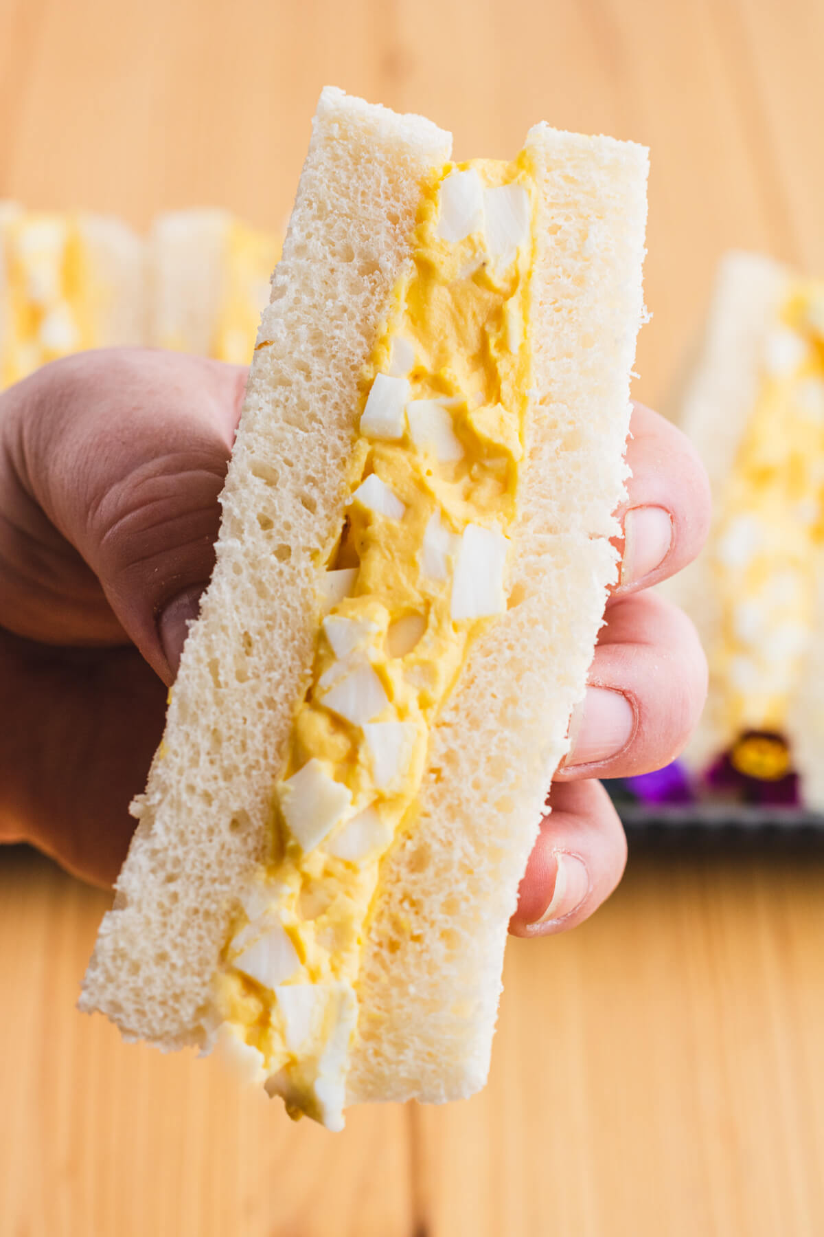 A hand holding a rich yellow egg salad sandwich on fluffy white crust less bread.