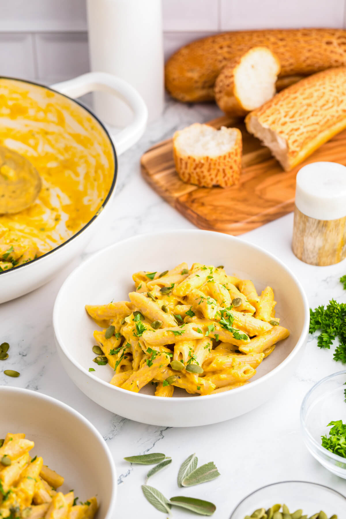 A table scene containing two white bowls filled with penne in pumpkin pasta sauce beside a sliced baguette