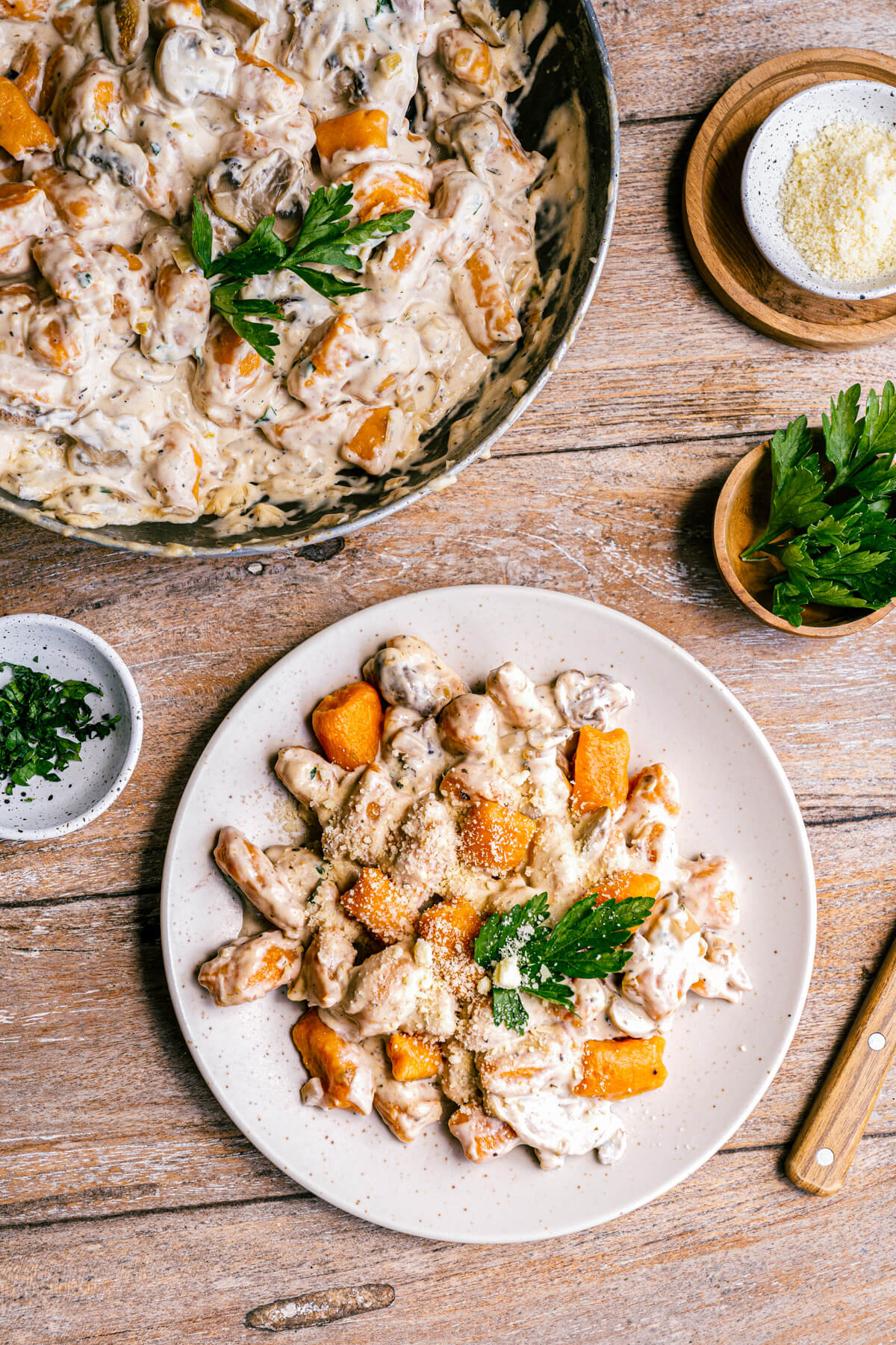 A table scene with a pan full of vibrant orange sweet potato gnocchi swimming in a creamy mushroom sauce beside a plate with a serving of creamy mushroom gnocchi.
