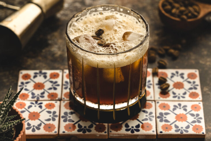 A foam topped nutty brown cocktail garnished with coffee beans in a rocks glass on Mexican patterned tiles.