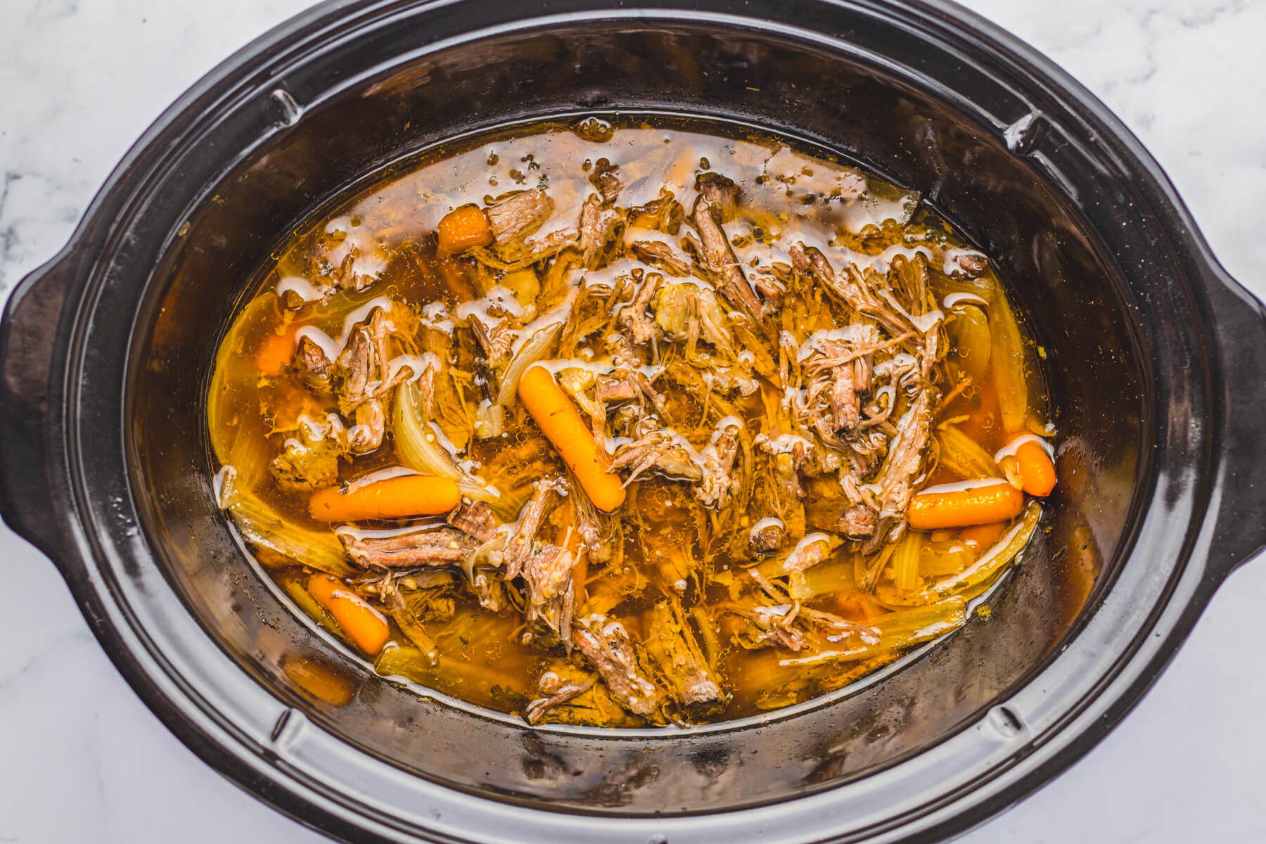 A black crock pot insert containing shredded chuck roast, carrots, and onions, in gravy.