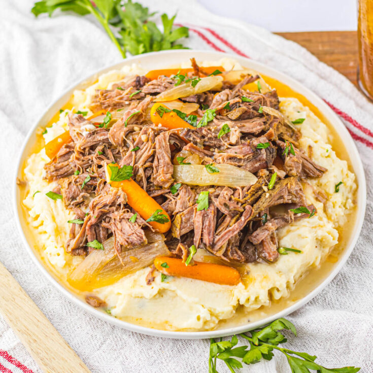 A plate full of shredded crock pot chuck roast, carrots, and onions on a bed of creamy mashed potatoes.