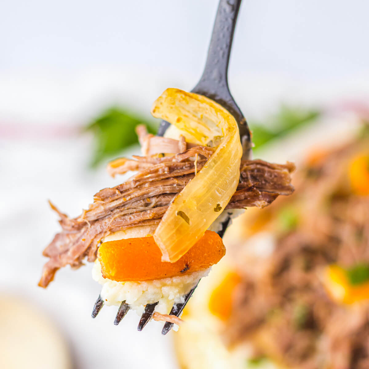 A fork holds a piece of tender chuck roast along with a baby carrot and golden caramelized onion.