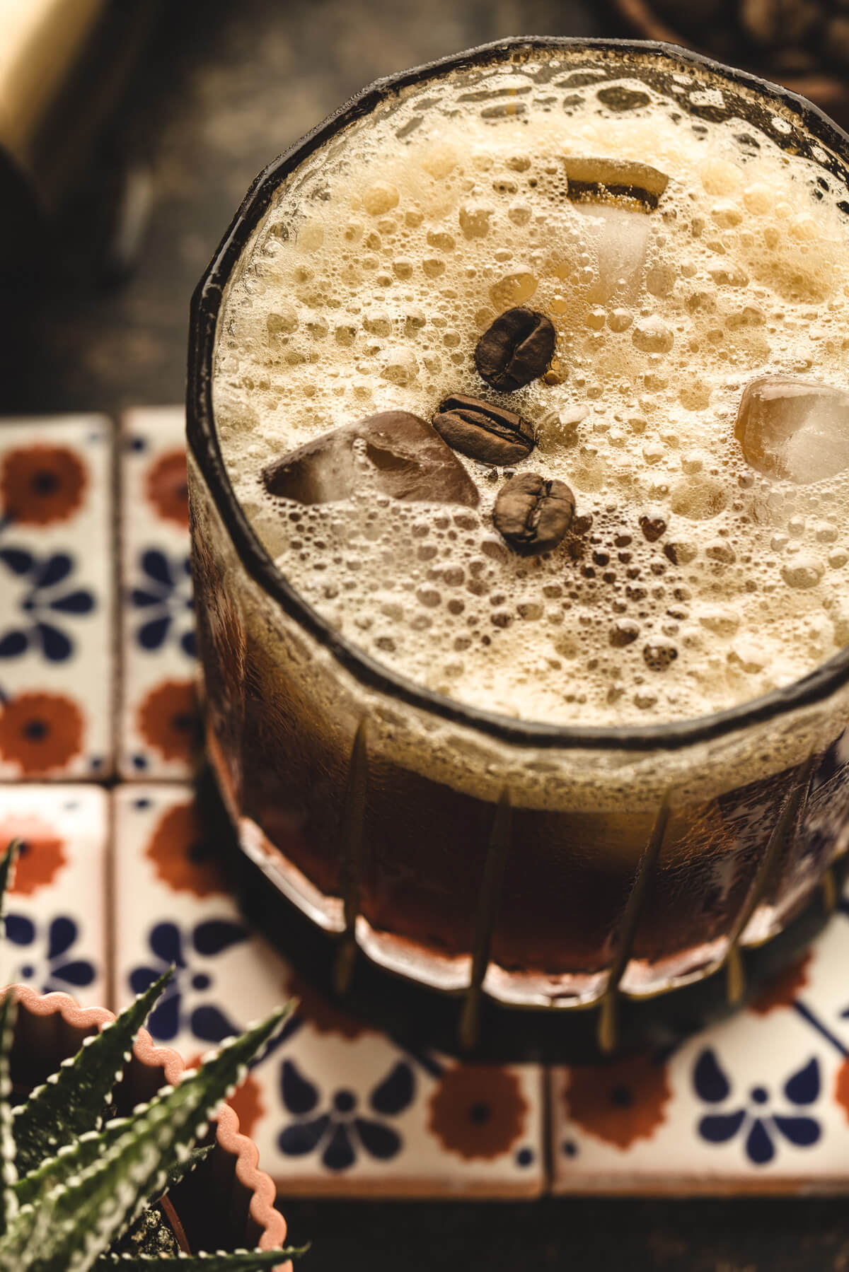 A foam topped nutty brown cocktail garnished with coffee beans in a rocks glass on Mexican patterned tiles. 