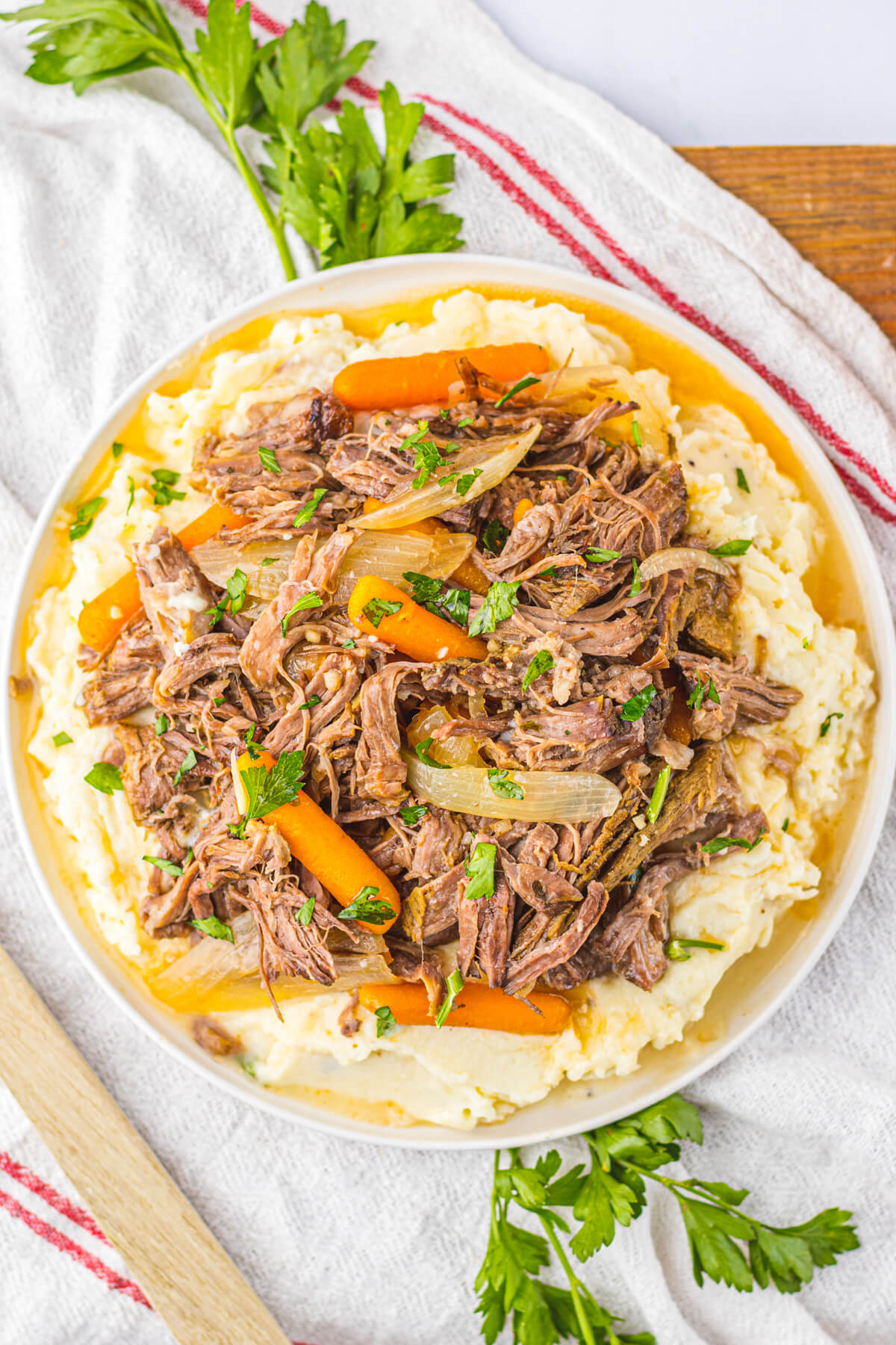 A plate full of shredded crock pot chuck roast, carrots, and onions on a bed of creamy mashed potatoes.