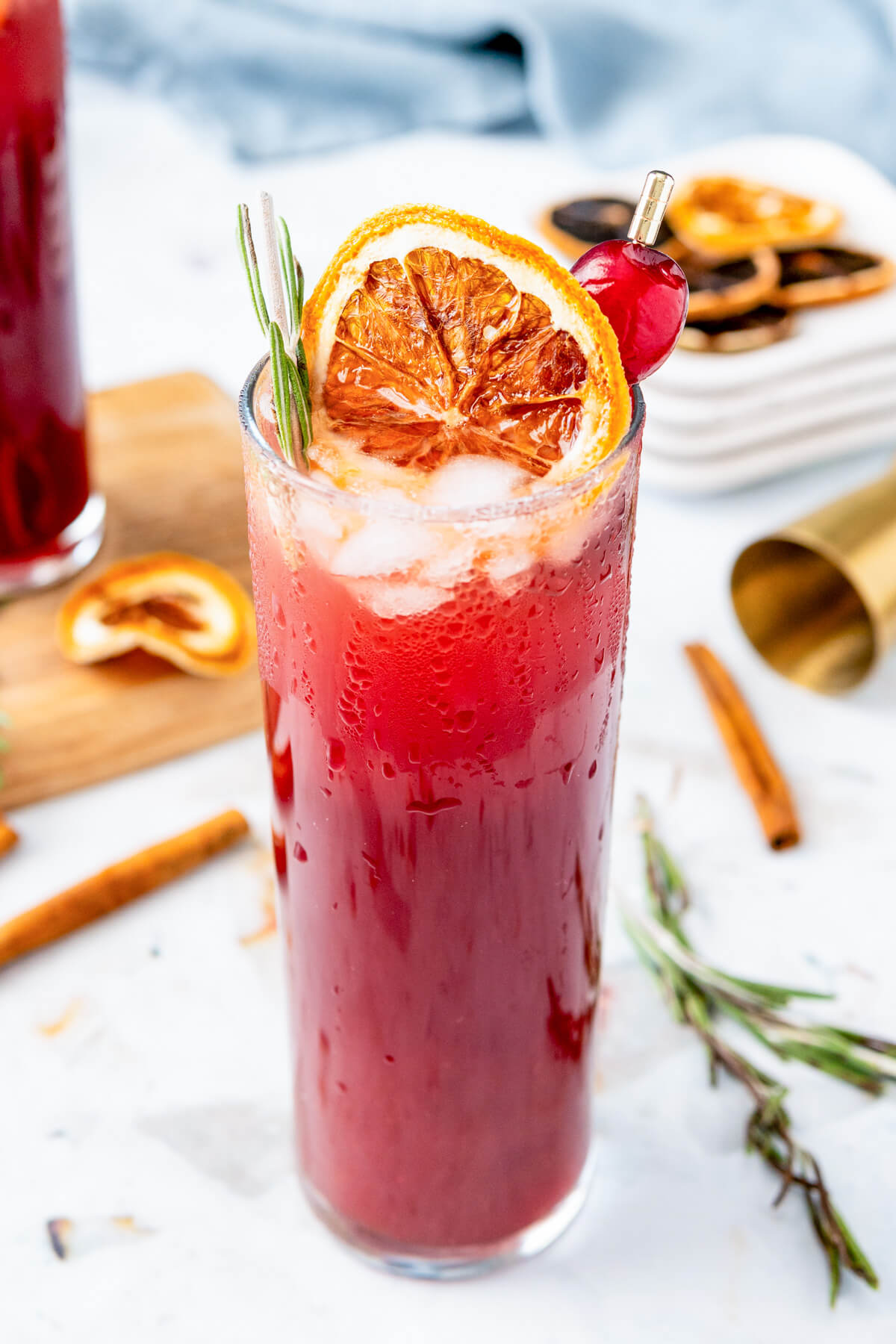 A highball cocktail glass filled with ruby red Mulled Cranberry Madras Cocktail garnished with with a dried orange wheel, cranberries, and a sprig of rosemary. 