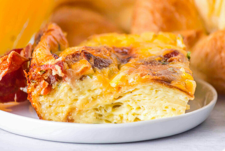 A square serving of Croissant breakfast casserole showing the layers of eggs, croissants, bacon, and cheese.