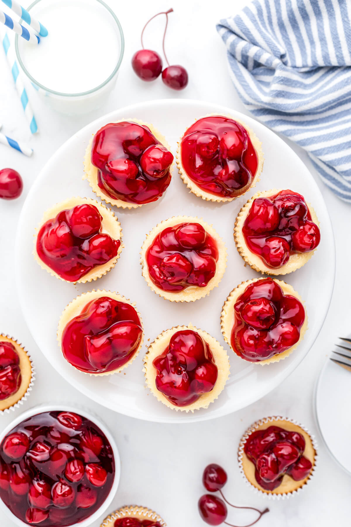 A group of Mini Cherry Cheesecakes on a white platter surrounded by cherry pie filling and fresh cherries.
