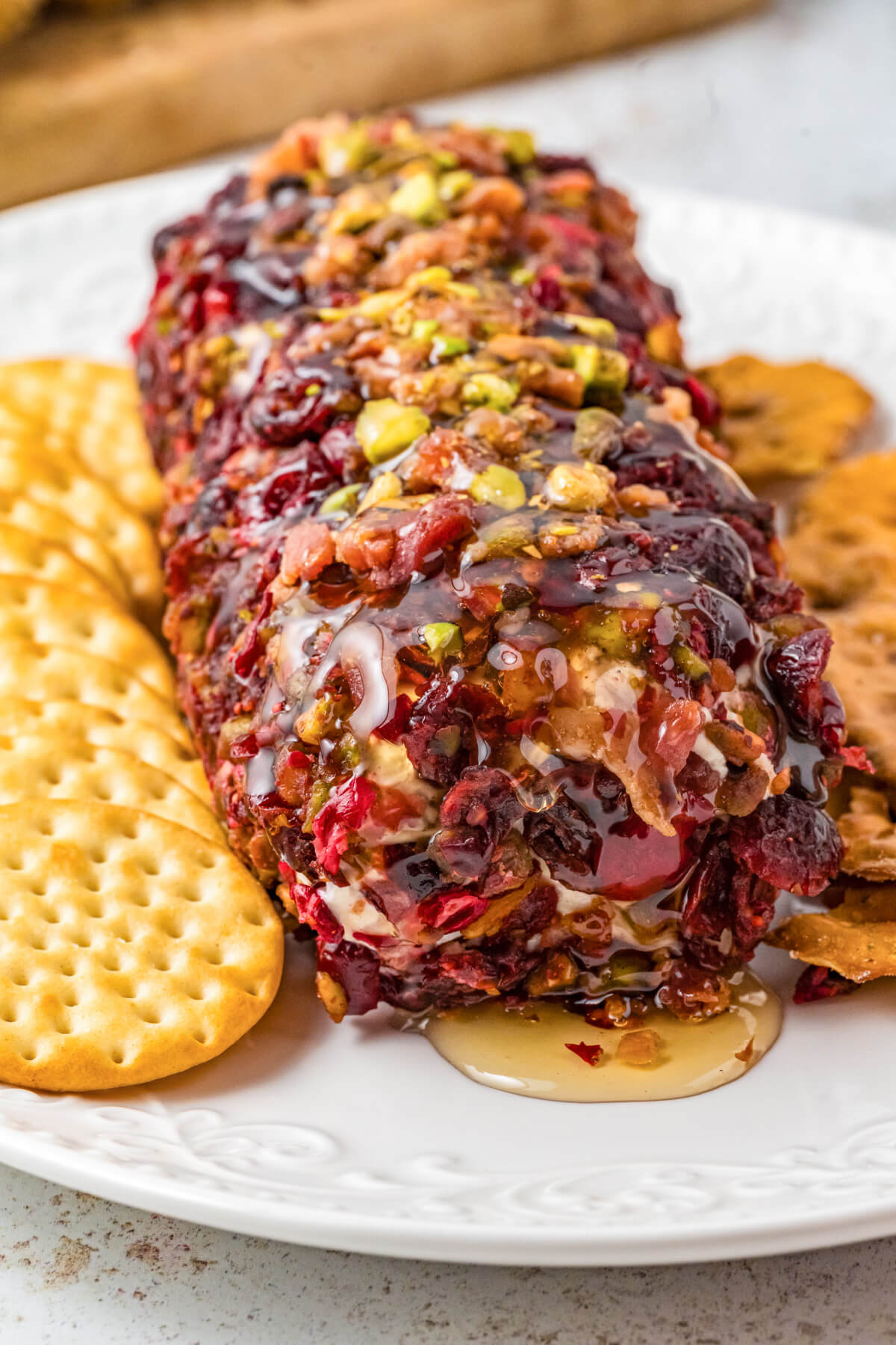 A whole honey coated pistachio and cranberry cheese log on a white plate surrounded by crackers.