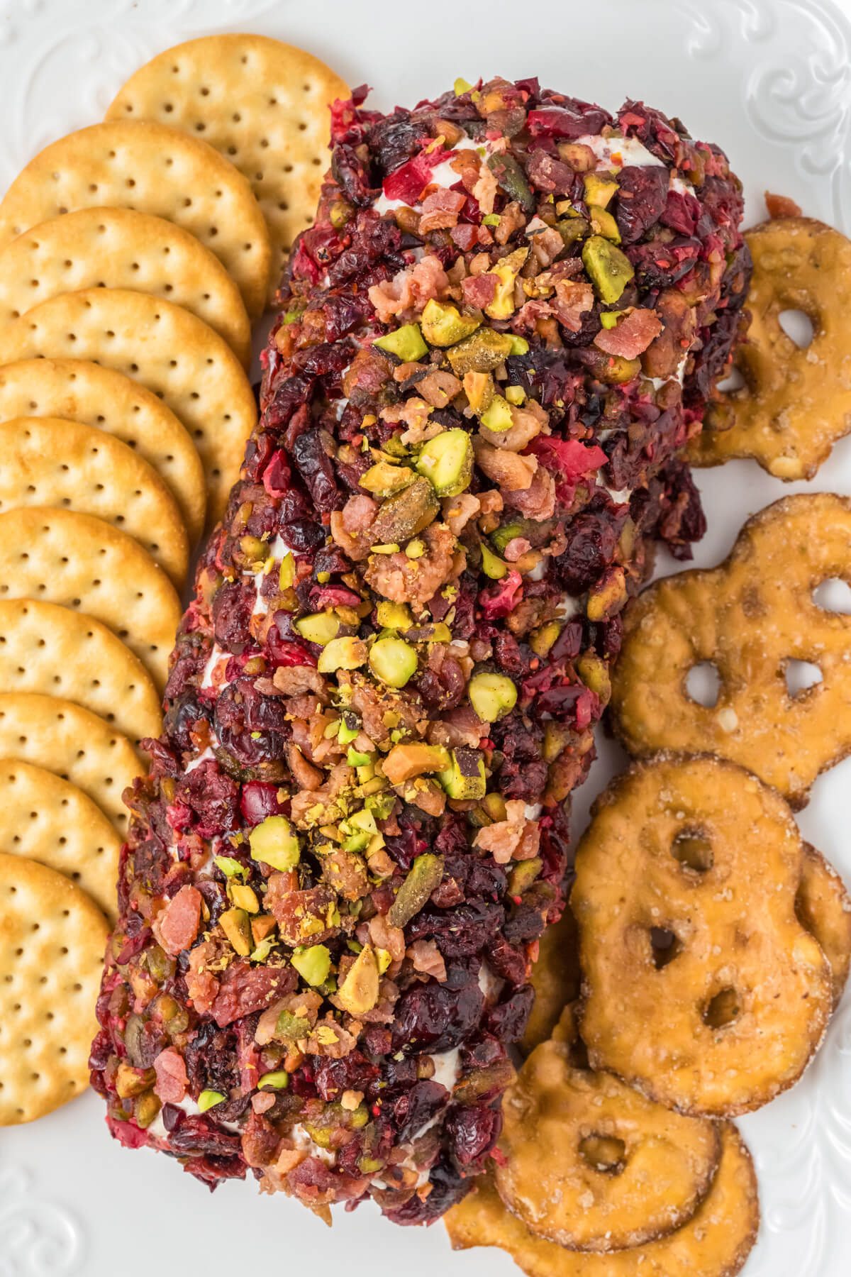 A whole honey coated pistachio and cranberry cheese log on a white plate surrounded by crackers.