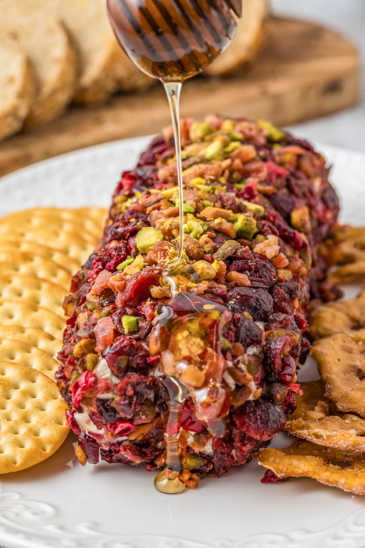 A whole pistachio and cranberry cheese log on a white plate surrounded by crackers being drizzled with golden honey.