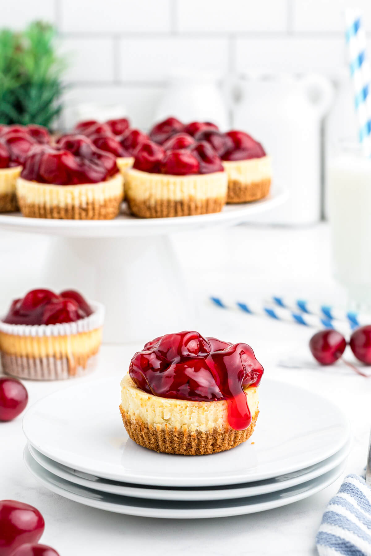 One mini Cherry Cheesecake with oozing topping sits on a stack of white plates with a platter of cheesecakes in background.