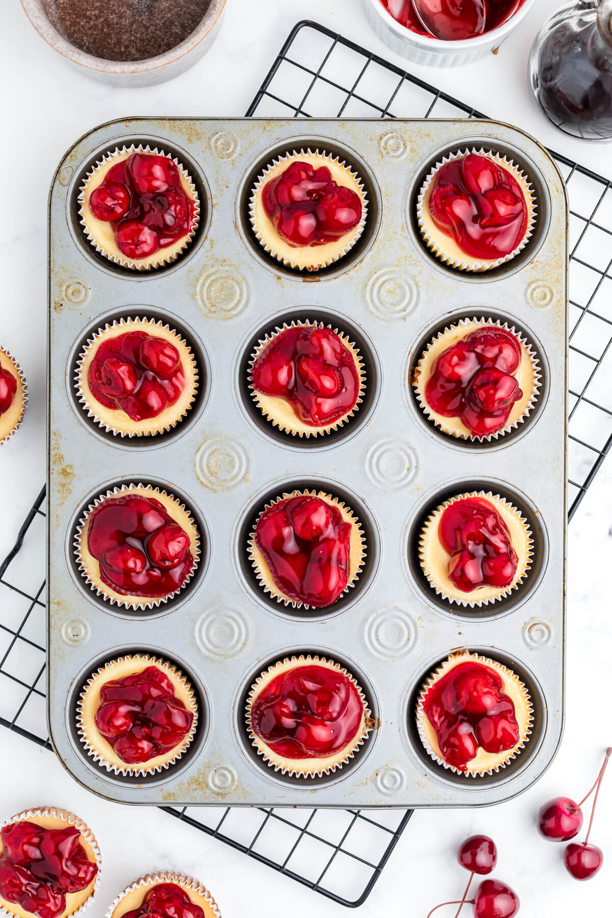 A muffin tin filled with mini cheesecakes topped with vibrant red cherry pie filling.