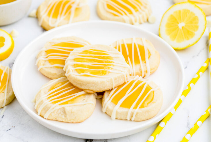 A group of Lemon curd thumbprint cookies topped with a white chocolate drizzle on a white plate.