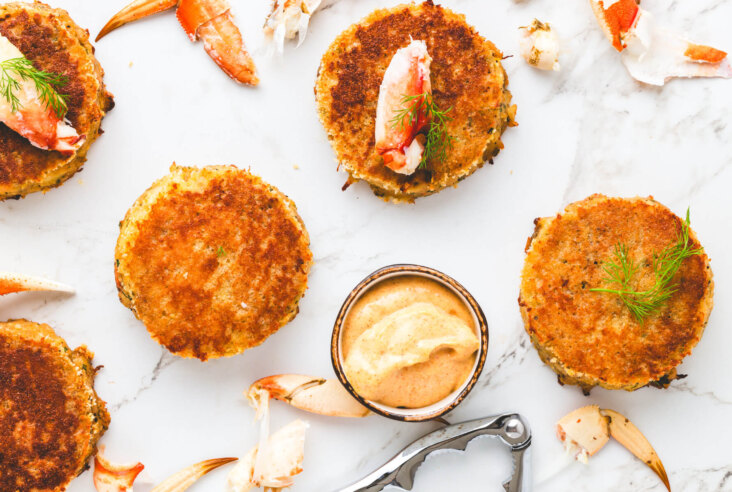 A table scene featuring several Old Bay Crab cakes, lump crab meat, garnishes and crab claws.