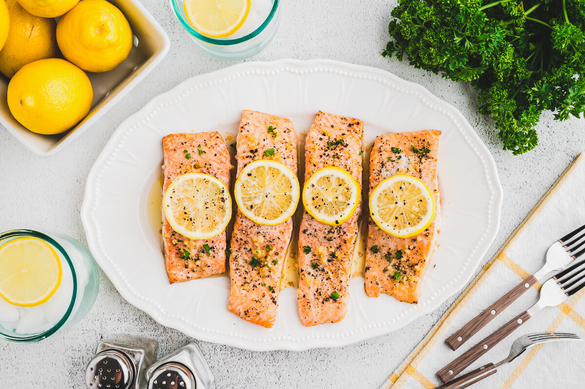 A white platter containing four oven baked lemon pepper salmon filets topped with lemon slices.