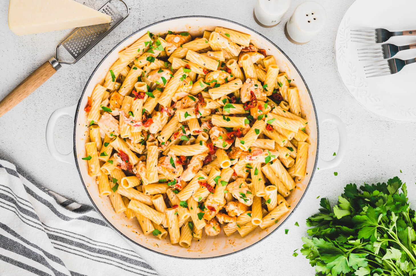 A table scene wit a white pan full of Marry Me Chicken Pasta featuring rigatoni pasta, chicken, sun dried tomatoes, and herbs in a creamy pasta sauce.