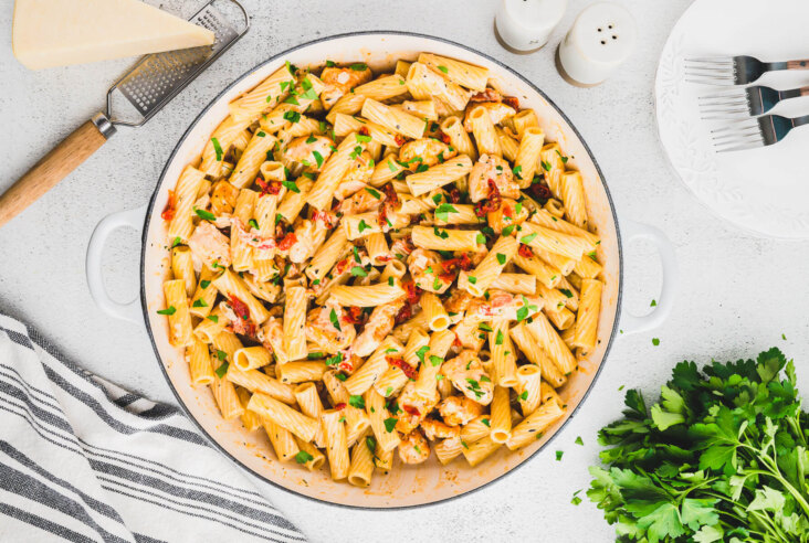 A table scene wit a white pan full of Marry Me Chicken Pasta featuring rigatoni pasta, chicken, sun dried tomatoes, and herbs in a creamy pasta sauce.
