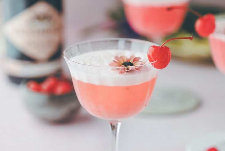 A pink lady cocktail garnished with a bright red cherry and an edible flower.
