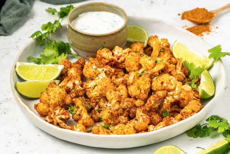 A plate of vibrant curried air fryer cauliflower beside a tiny bowl of creamy dip, cilantro, and lime wedges.
