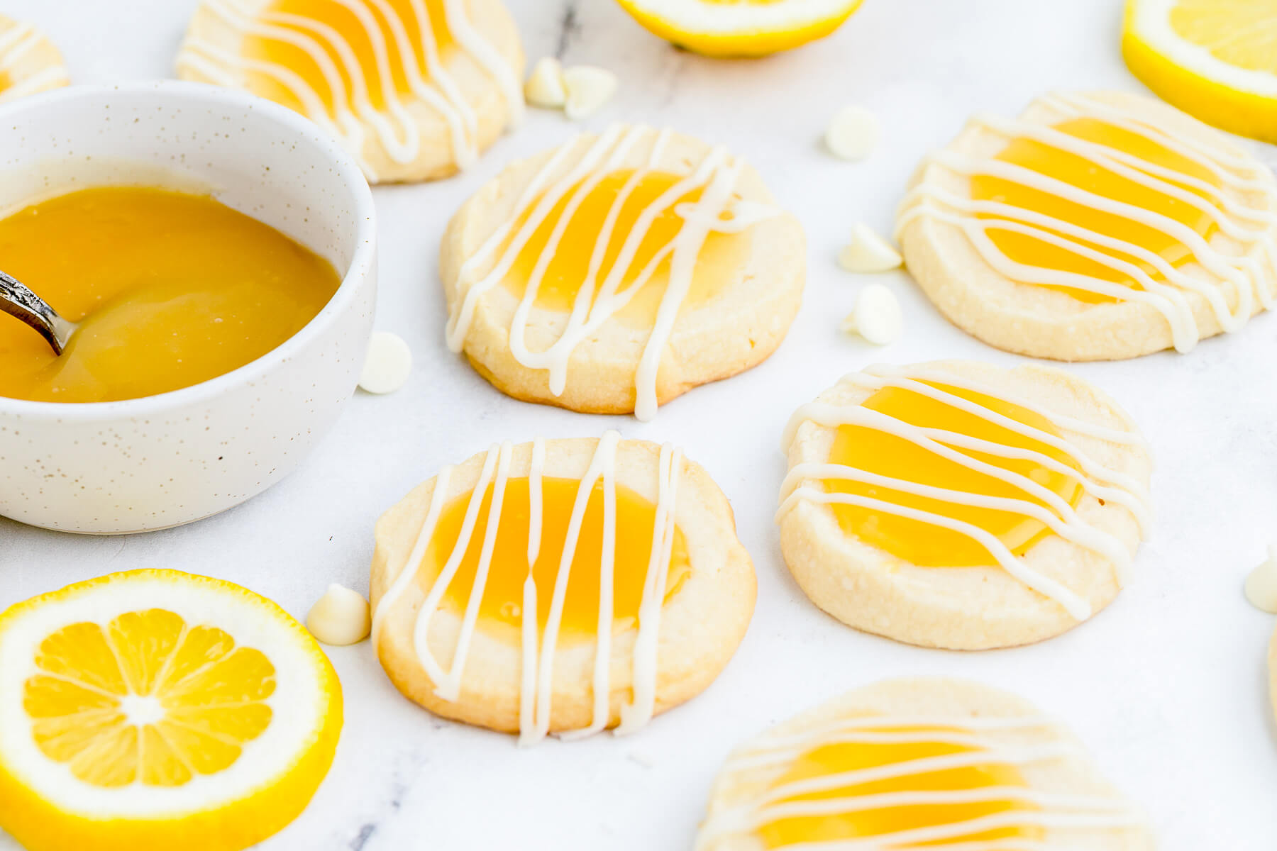 A group of Lemon curd thumbprint cookies topped with a white chocolate drizzle with a small bowl of lemon curd on white marble.