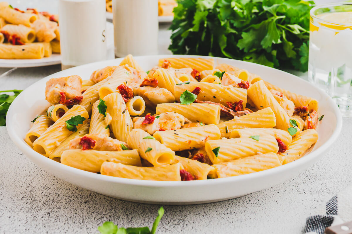 A white bowl of Marry Me Chicken Pasta featuring rigatoni pasta, chicken, sun dried tomatoes, and herbs in a creamy pasta sauce.