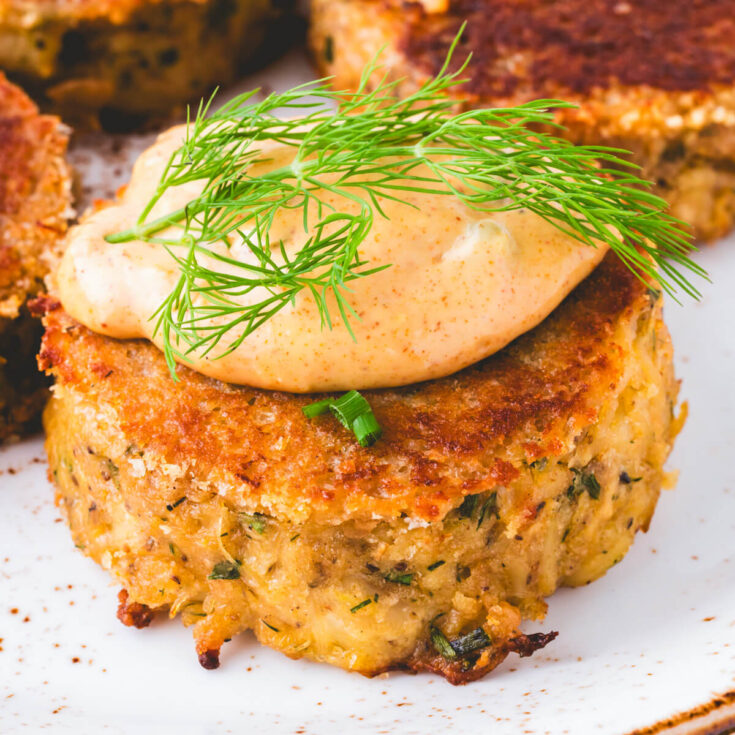 A golden fried crab cake on a white plate with a dollop of Cajun aioli and fresh dill.