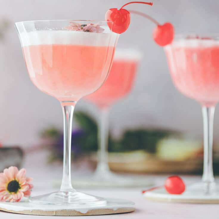 Three coupe glasses garnished with red stemmed cherries filled with foamy pink lady cocktail.