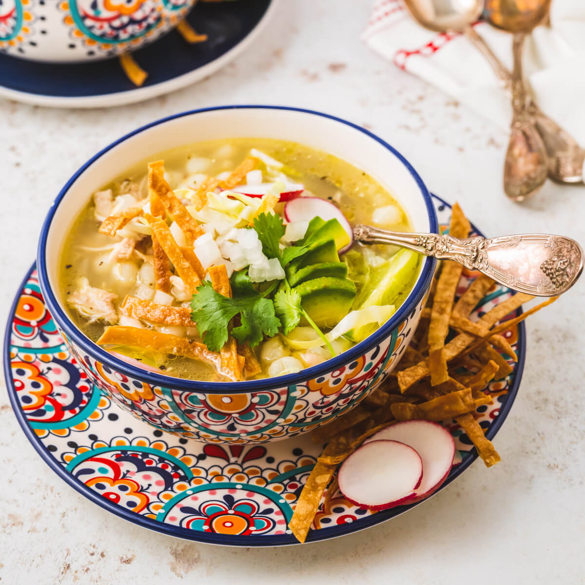 A colourful soup bowl containing Pozole blanco featuring white hominy, cilantro, radishes, cabbage, lime wedge, and strips of fried tortilla.