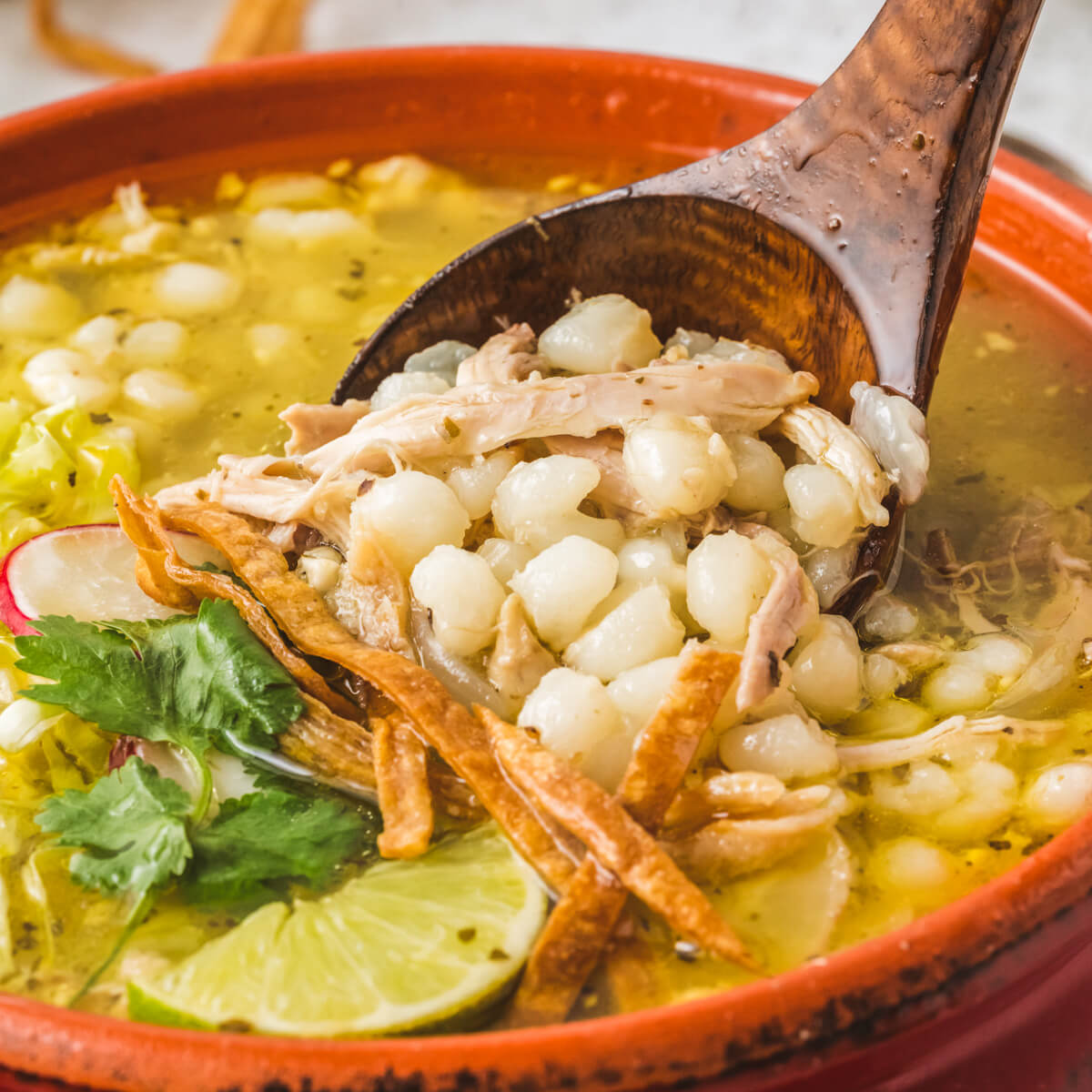 A wooden spoon in bright terra cotta pot filled with Pozole blanco featuring white hominy, cilantro, radishes, cabbage, lime wedge, and strips of fried tortilla.