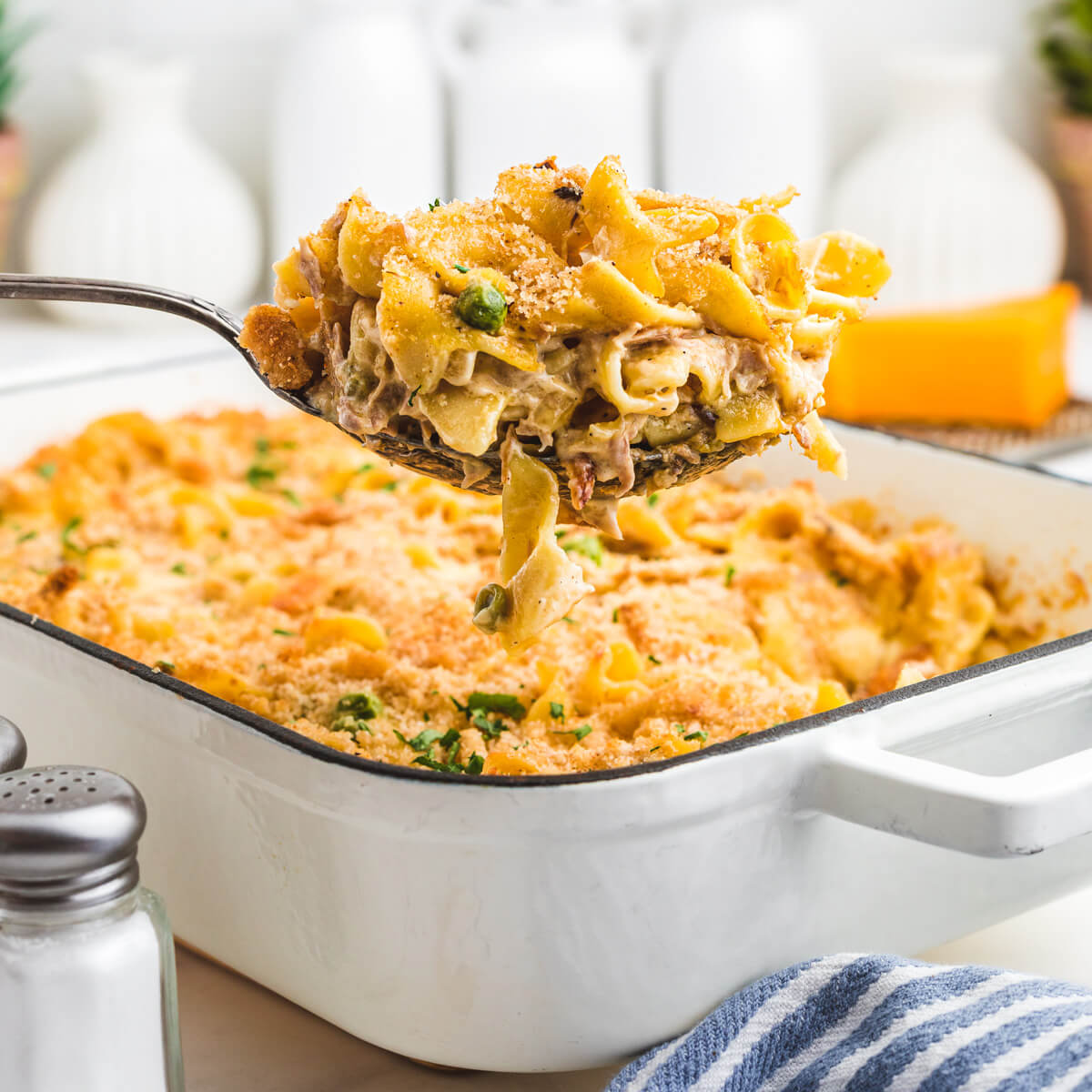 A serving spoon containing a serving of tuna noodle casserole hovers over a baking dish. 